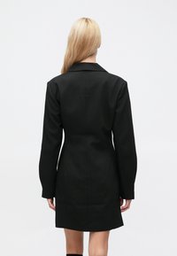 Černé šité sako ve tvaru šatů s výraznou siluetou, velkými klopami, dlouhými rukávy a hladkou texturou látky.