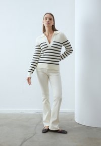 Striped v-neck sweater in cream and black with long sleeves, paired with white flared pants and black flats, on a concrete floor.