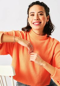 Ribbet, højhalset sweater i livlig orange. Modellen bærer sølvarmbånd og ringøre. Baggrunden er neutral for at fremhæve outfittet.