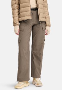 Persoon in een beige pufferjack, taupe cargobroek met klepzakken, vingervrije handschoenen en beige sportschoenen op een witte achtergrond.