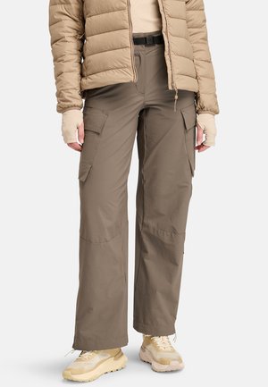 Persona vistiendo una chaqueta acolchada beige, pantalones cargo topo con bolsillos de solapa, guantes sin dedos y zapatillas deportivas beige sobre un fondo blanco.