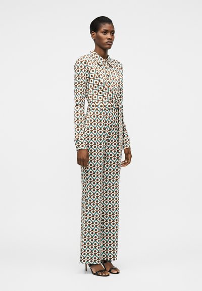 Γυναίκα στέκεται με ολόσωμη φόρμα μακρυμάνικη που έχει γεωμετρικό μοτίβο διαμαντιού και κύκλων σε καφέ, άσπρο και τυρκουάζ, συνδυασμένη με μαύρες γόβες.