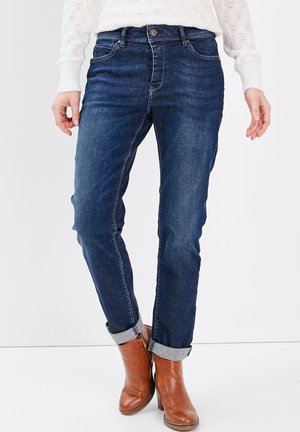 Jean en denim foncé avec une coupe slim, taille mi-haute, style à cinq poches et ourlets retroussés, associés à des bottines en cuir marron.