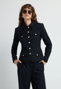 Czarna marynarka dopasowana z złotymi guzikami i czterema przednimi kieszeniami, z kołnierzowym dekoltem i wyraźną sylwetką. W zestawie z czarnymi spodniami.