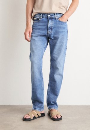 Svetlo modri jeans s ravnim krojem, pet žepov in klasičnim gumbnim zapiranjem, v kombinaciji s svetlečimi sandali v barvi tan.