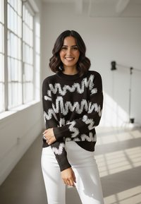 Brązowy sweter dziany z białym wzorem w zygzak, z okrągłym dekoltem i długimi rękawami, zestawiony z białymi spodniami w jasnym, dobrze oświetlonym pomieszczeniu.