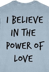 T-shirt en coton bleu clair avec un grand texte noir au dos : « JE CROIS EN LE POUVOIR DE L'AMOUR » dans un style manuscrit en gras.