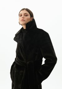 Černý kabát z umělé kožešiny s vysokým límcem, dvojitým zapínáním, pásem v pase a bočními kapsami, s měkkým a strukturovaným povrchem.