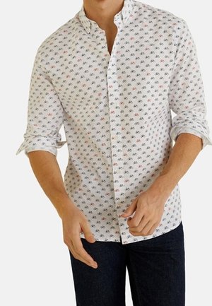 Homme portant une chemise blanche à boutons avec un petit motif de vélos rouges et noirs, manches retroussées, associée à un pantalon sombre.