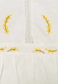 Vitt tyg med broderi av gula och bruna löv. Har en spetsinsats och veckad fåll längst ner.