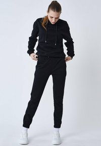 Zwarte hoodie met trekkoorden en reliëflogo, gecombineerd met bijpassende zwarte joggers. Witte sneakers maken de look compleet. Soepele stofstructuur.