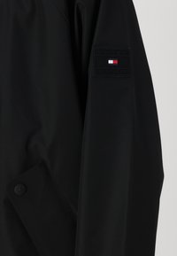 Czarna kurtka wykonana z teksturowanego materiału, z zapięciem na guziki i naszywką z logo na rękawie w kolorach czerwonym, białym i niebieskim.