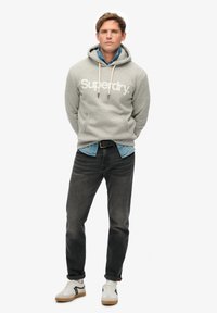 Un sweat à capuche gris avec le logo "Superdry" blanc, associé à une chemise en jean clair, un jean foncé et des baskets blanches avec des accents noirs et beiges.