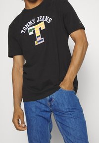 Svart bomullströja med en färgglad "T"-logotyp och texten "TOMMY JEANS". Kortärmad, avslappnad passform, kombinerad med blå denimjeans.