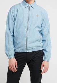 Ljusblå jeansjacka med dragkedja fram, knappad krage, elastiska ärmmuddar och två sidofickor. Har en liten logotyp på bröstet.