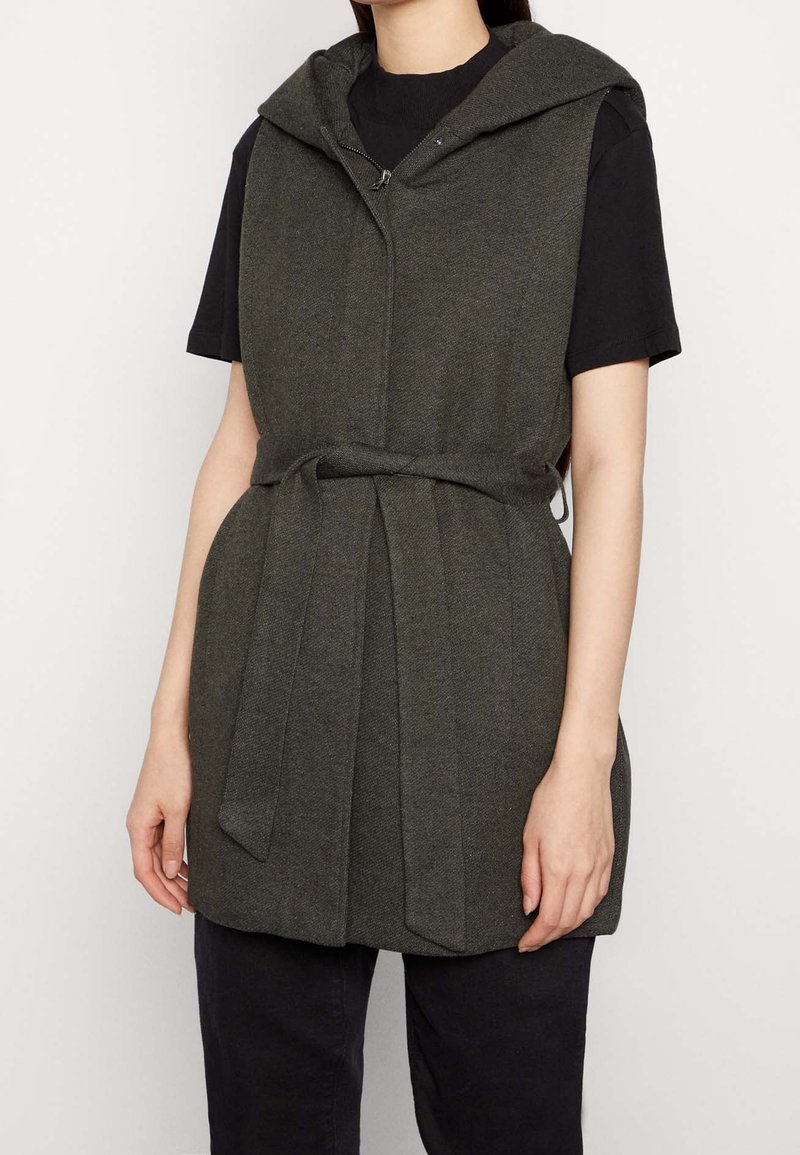 Ärmellose Kapuzenweste aus dunkelgrünem Stoff, mit Reißverschluss vorne, tailliertem Gürtel und strukturierter Oberfläche. Kurze Ärmel unter der Weste.