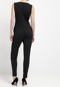 Combinaison noire sans manches avec une silhouette ajustée, une texture lisse et une fermeture éclair discrète au dos, complétée par des jambes fuselées.