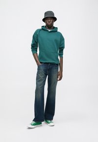 Turkusowa bluza z kapturem z kieszenią z przodu, niebieskie szerokie dżinsy i zielono-białe trampki, uzupełnione czarnym kapeluszem typu bucket.