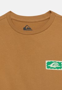 Καφέ μπλουζάκι με στρογγυλή λαιμόκοψη, με μικρό πράσινο και λευκό λογότυπο στο στήθος και εκτυπωμένο λογότυπο της μάρκας μέσα στον γιακά.