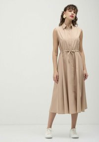 Abito beige senza maniche con bottoni frontali, vita regolabile con cordoncino e orlo fluente. Indossato con sneakers bianche. Tessuto morbido e design pulito.