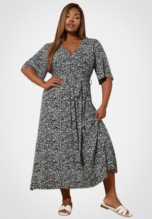 Kvinde iført en sort og hvid blomsterprint slå-om kjole med albuelange ærmer og hvide slidesandaler, poserer mod en ensfarvet baggrund.