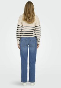 Stickad tröja i krämfärg med marinblå ränder, i kombination med raka blå jeans. Synliga fickor på jeansen och vita sneakers. Bakifrån.