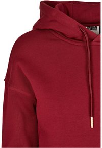 Sudadera de algodón roja con capucha ajustable mediante cordones, puños acanalados y corte holgado. La textura es suave con detalles de costuras visibles.