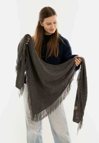 Grijze gebreide sjaal met franjes, lichte textuur en een driehoekige vorm, vastgehouden door iemand die een marineblauwe top draagt.