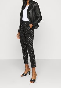 Černé kalhoty s potiskem bílých puntíků, těsně přiléhající, v kombinaci s bílým tričkem a černou koženou bundou. Celkový vzhled doplňují černé sandály na otevřené špičce.