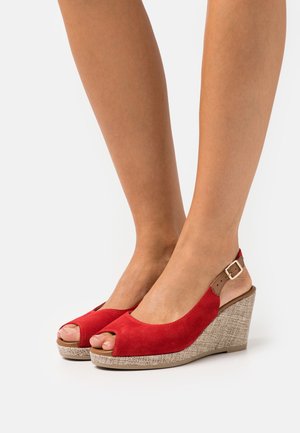 Legs wearing red suede open-toe wedge sandals with beige textured heels and ankle straps on a white background.