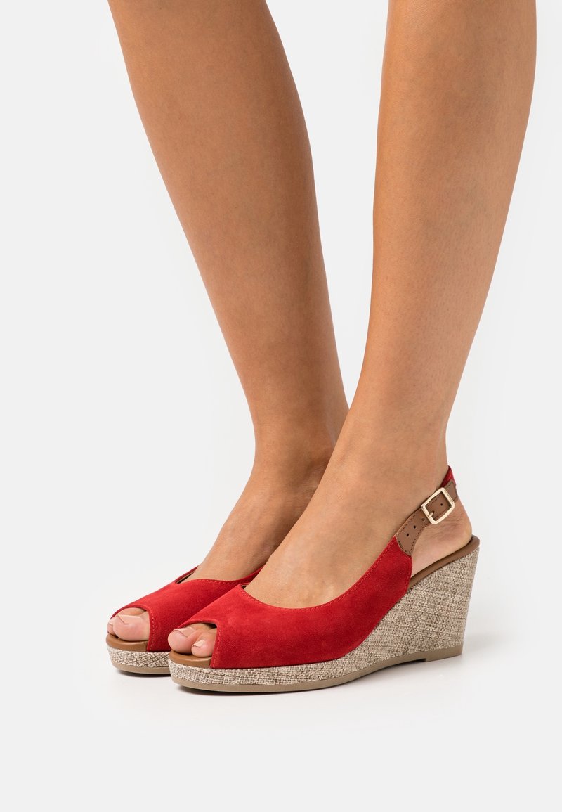 Jambes portant des sandales à talons compensés ouvertes en daim rouge avec des talons texturés beiges et des sangles autour des chevilles sur un fond blanc.