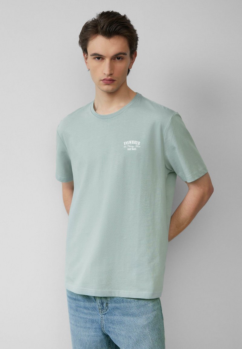 Een jonge man met bruin haar, gekleed in een lichtgroen T-shirt met korte mouwen en witte tekst, en een blauwe spijkerbroek. Hij staat tegen een effen lichte achtergrond.