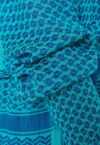Tissu turquoise avec des motifs géométriques et floraux bleu foncé, montrant la texture et les plis sur un vêtement à manches longues.