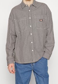Camicia a righe in bianco e nero, con due tasche sul petto, colletto classico e vestibilità ampia. Il tessuto sembra avere una texture.