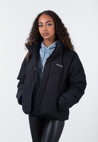 Schwarze Daunenjacke mit hohem Kragen, Frontreißverschluss und Seitentaschen. Verfügt über ein Carhartt-Logo auf der Brust und eine gesteppten Textur.