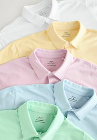 Fünf Poloshirts in einem Stapel präsentiert: weiß, pastellgelb, pastellrosa, hellblau und mintgrün. Regelmäßige Passform, Baumwollmaterial, Knopfkragen.