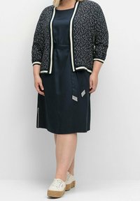 Abito blu navy con finitura in satin e dettagli a cravatta, abbinato a un cardigan a motivi bianco e nero. Sneakers bianche con piattaforma completano il look.