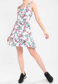 Blommig klänning med vit bas, prydd med gröna blad och rosa blommor. ärmlös design, utställd silhuett, ihop med svarta sneakers.