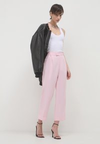 Donna che indossa pantaloni a vita alta di un rosa chiaro, una canotta bianca, sandali neri con tacco e tiene una giacca di pelle nera su una spalla.