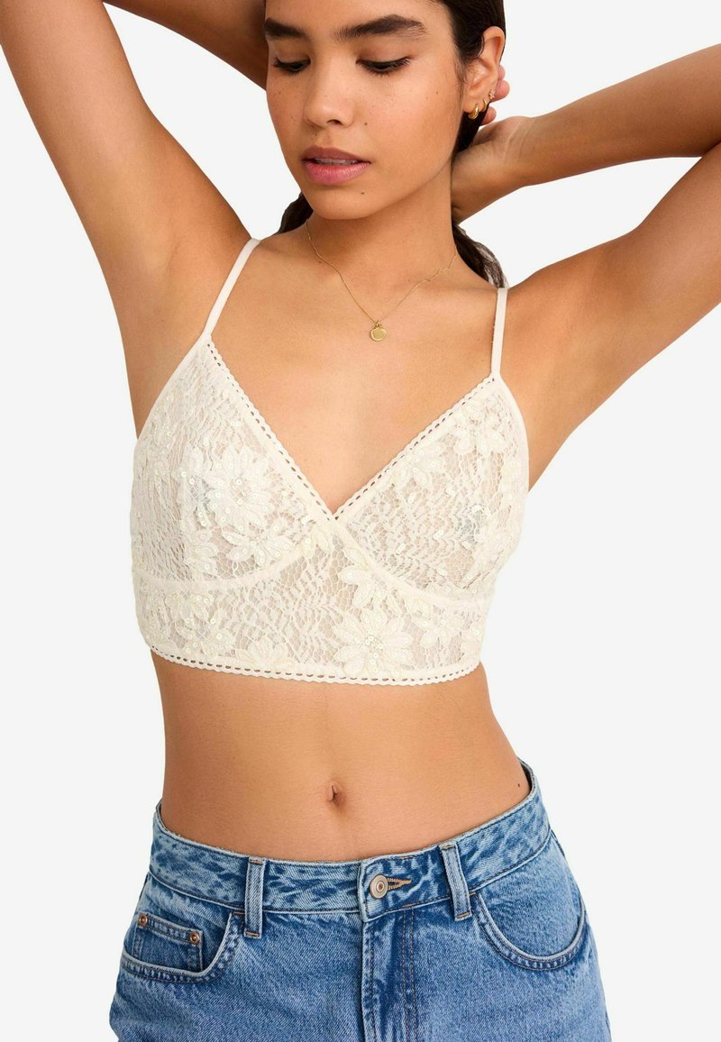 Romantische kanten bralette in crème met een bloemmotief, doorschijnende textuur, V-halslijn, verstelbare bandjes en geschulpte rand, gecombineerd met high-waisted denim shorts.