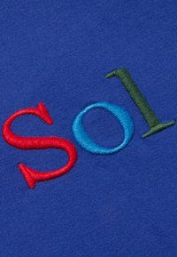 Modrá látka s vyšitými písmeny "S", "o", "l" a "1" v červené, modré a zelené barvě, s hladkou texturou a kontrastními barvami.