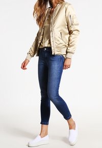 Giacca bomber color oro metallizzato con chiusura a zip, tasche laterali e polsini a costine, abbinata a jeans skinny blu e scarpe slip-on bianche.