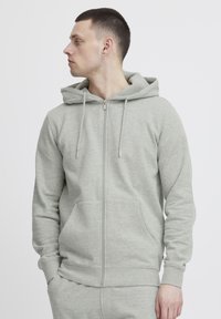 Felpa zip grigia chiara realizzata in morbido tessuto, dotata di cappuccio con cordino, tasche frontali e polsini a coste. Design semplice e senza cuciture.