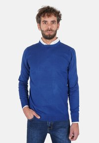 Maglione blu lavorato a maglia con scollo rotondo, polsini e orlo a coste. Abbinato a una camicia bianca con colletto sotto. Indossato con jeans scuri.