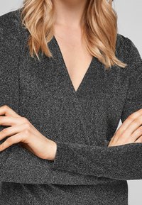 Svart, texturerad tygblus med djup V-ringning, lång ärm och en subtil glans. Har en omslagsdesign i midjan som framhäver silhuetten.