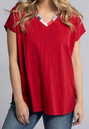 Vrouw draagt een loszittend rood T-shirt met korte mouwen en V-hals met meerkleurige biezen, gecombineerd met een blauwe spijkerbroek, staand tegen een effen achtergrond.