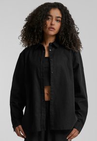 Black button-up shirt made of a smooth fabric, featuring a pointed collar and long sleeves, worn over a black cropped top.