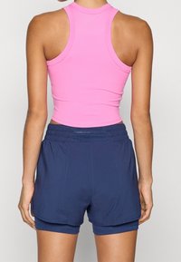Femme de dos portant un crop top rose sans manches et un short de sport bleu marine avec une couche intérieure serrée sur un fond uni.
