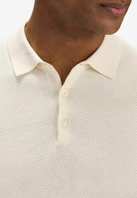 Krämfärgad polo med en texturerad, bikupemönster. Mjukt tyg, treknappsknäppning och en klassisk kragsdesign.