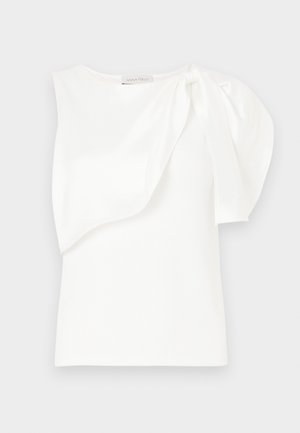 Top blanco sin mangas con detalle de tela en capas asimétricas y manga abullonada en un hombro, dispuesta sobre un fondo blanco.